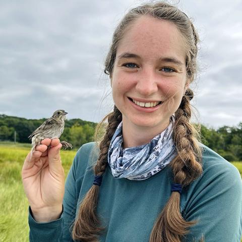 Photo of student holding a bird in her fingers outside. 