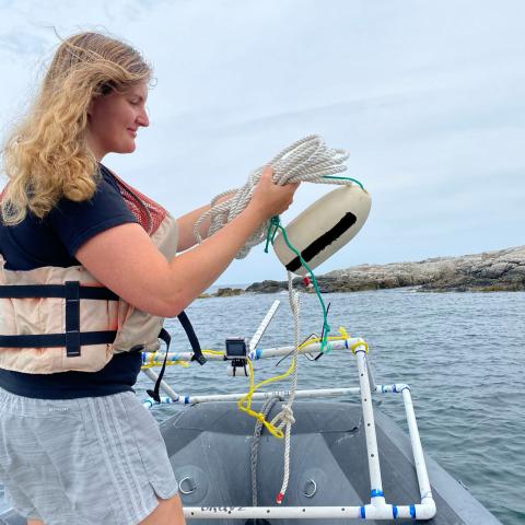 Photo of student on boat holding instrumentation by rocky shoreline. 