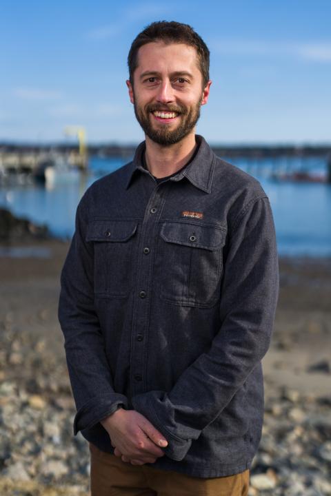 Linas Kenter, a white man with short brown hair and beard, stands on a rocky beach wearing a grey button down shirt.